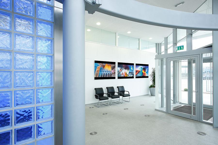 Empfang Ruhiger Empfangsbereich mit Glaswand und mehreren Sitzplätzen. An der Wand hängen drei Bilder mit lebhaften Farben. Die Eingangstür ist aus Glas, und in der Ecke steht eine Pflanze.