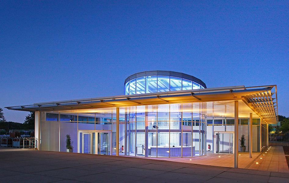 Neubau eines Empfangsgebaeudes Moderne Architektur eines Gebäudes bei Dämmerung. Die Fassade ist aus Glas und Metall, mit einem runden Dach. Sanfte Beleuchtung hebt die Struktur hervor und schafft eine einladende Atmosphäre. Um das Gebäude herum sind Pflanzen und eine gepflasterte Fläche sichtbar.