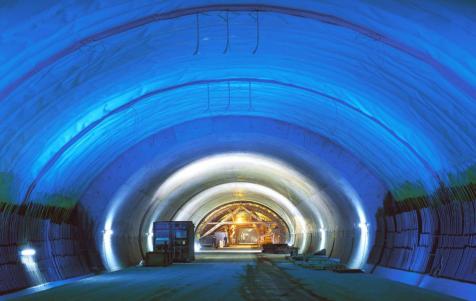 Autobahntunnel A81 im Bau Ein beleuchteter Tunnel mit blauen Wänden und einem gewölbten Dach. Im Hintergrund sind einige Baugeräte und Personen zu sehen. Der Boden ist eben und der Tunnel scheint gut strukturiert zu sein.