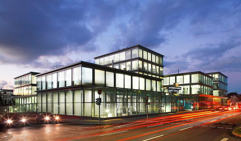 Schwäbische Zeitung Redaktionsgebäude Moderne Bürogebäude mit Glasfassade bei Nacht, beleuchtet von Innen. Straßenverkehr ist sichtbar, mit Lichtspuren von fahrenden Autos. Der Himmel ist bewölkt und es ist Dämmerung.