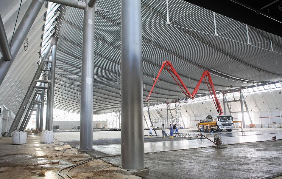 Baustelle Landesmesse Stuttgart Ein großer, leerer Innenraum mit hohen, metallic schimmernden Wänden und einem gewölbten Dach. In der Mitte befindet sich ein Betonmischer mit einem langen Schlauch, während Arbeiter auf dem Boden beschäftigt sind. Stützen aus Edelstahl sind sichtbar.