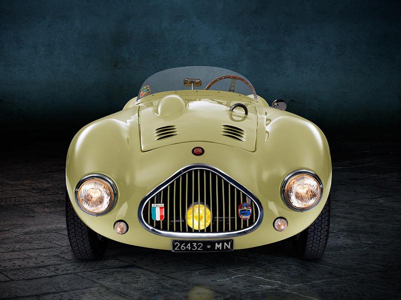 FIAT 500 Graziani MM Frontal Ein klassisches, gelbgrünes Sportauto mit runder Form und zwei schmalen Scheinwerfern steht auf einem grauen, gepflasterten Boden. Im Hintergrund ist eine dunkle Wand sichtbar, die eine elegante Atmosphäre schafft.