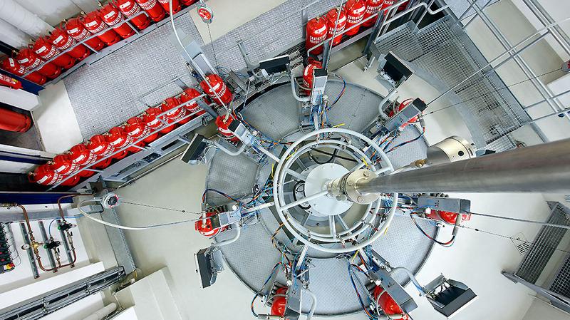 Abfuellanlage Eine Vogelperspektive auf eine kreisförmige Anordnung von technischen Geräten und Rohren. Um den zentralen Bereich sind mehrere rote Gasflaschen auf einer geraden Plattform angeordnet. Der Boden ist grau und die Umgebung wirkt industriell.