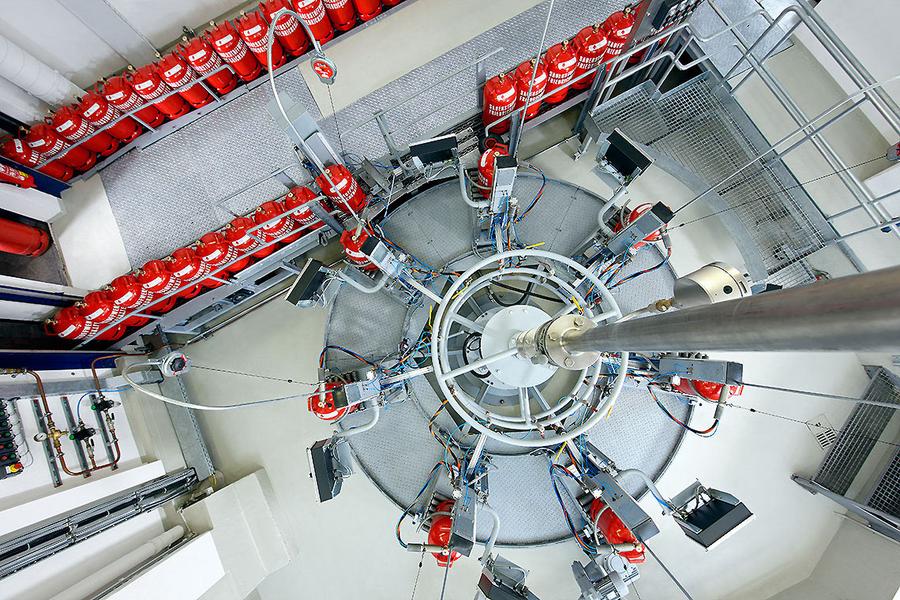 Abfuellanlage Eine Vogelperspektive auf eine kreisförmige Anordnung von technischen Geräten und Rohren. Um den zentralen Bereich sind mehrere rote Gasflaschen auf einer geraden Plattform angeordnet. Der Boden ist grau und die Umgebung wirkt industriell.