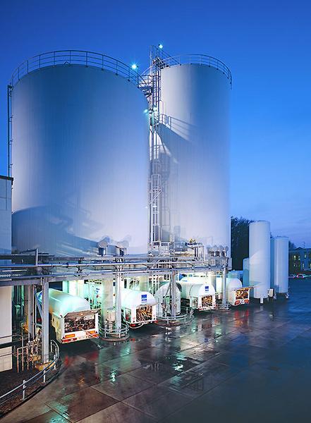 LKW Beladung Zwei große weiße Silos sind bei Dämmerung zu sehen, umgeben von einer glatten, nassen Fläche. In der Nähe befinden sich mehrere Tanks, die in Betrieb sind, und LEDs leuchten in Blau. Die Atmosphäre wirkt industriell und modern.