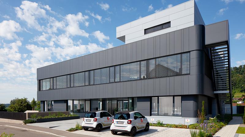Schmieder Gebaeude Modernes, mehrstöckiges Gebäude mit großen Fenstern und grauer Fassadenverkleidung. Vor dem Gebäude stehen zwei kleine Autos auf einem gepflasterten Parkplatz. Im Hintergrund sind Bäume und ein blauer Himmel mit einigen Wolken sichtbar.