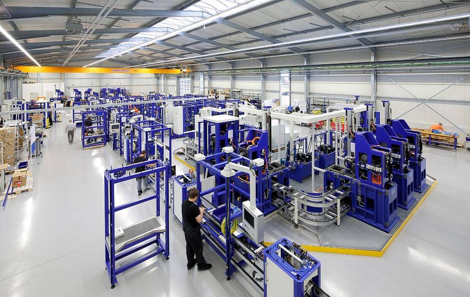 Testaufbau Fertigungsanlage Eine weite Aufnahme einer modernen Produktionshalle, die mit verschiedenen Maschinen und Fertigungsstationen in Blau und Weiß ausgestattet ist. Arbeiter sind in der Halle aktiv. Der Raum hat hohe Decken und viel natürliches Licht, das durch große Fenster strömt.