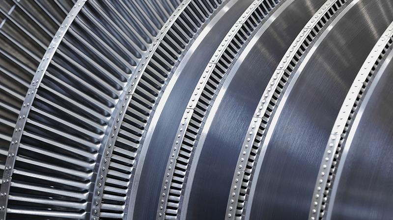 Dampfturbine Nahaufnahme von mehreren metallenen Turbinenschaufeln, die spiralförmig angeordnet sind. Die Oberfläche ist glänzend und zeigt feine Rillen sowie präzise Befestigungsdetails. Die Struktur deutet auf eine komplexe technische Konstruktion hin.