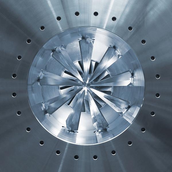 Verdichter Ein runder metallischer Gegenstand mit einer spiralförmigen Anordnung von scharfen Klingen in der Mitte. Der Hintergrund ist silbern und glänzend, mit kleinen Löchern, die gleichmäßig um die Klingen herum angeordnet sind.