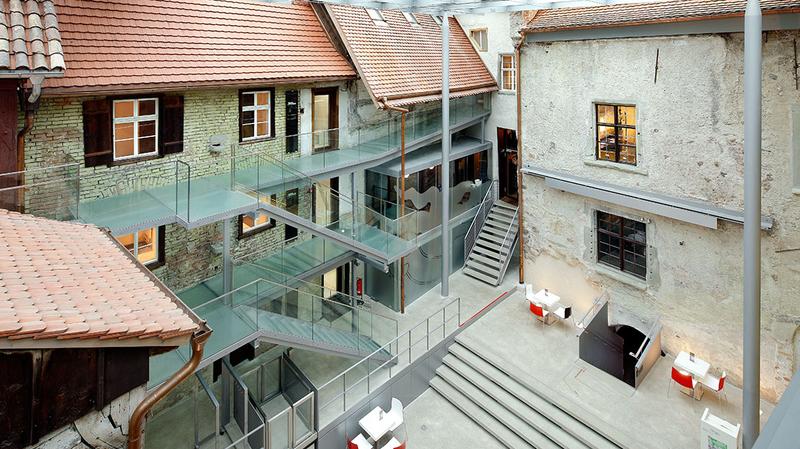 Innenansicht Humpis Museum Ein Überblick über ein modernes Atrium mit transparenten Glasböden und einer Kombination aus historischen Gebäuden. Die Umgebung ist hell erleuchtet, mit Treppen und Sitzbereichen. Die Architektur verbindet Alt und Neu in harmonischer Weise.