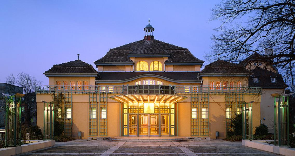 Festsaal in Leimen bei Heidelberg Historisches Gebäude mit stilvollem Eingang, beleuchtet in der Dämmerung. Die Fassade zeigt eine Mischung aus traditioneller und moderner Architektur, umgeben von Bäumen und einem geräumigen Vorplatz.