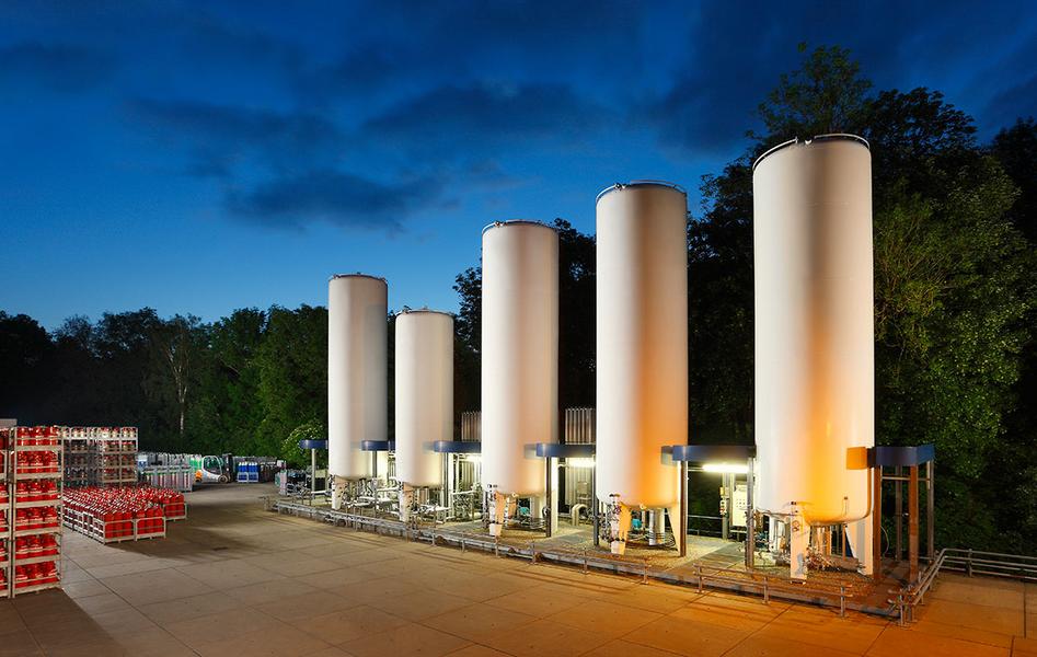 Tankanlage Fünf große, weiße Zylinderspeicher stehen in einer Außenanlage, umgeben von Bäumen. Im Hintergrund sind einige Paletten oder Kästen zu sehen. Die Szene ist bei Dämmerlicht aufgenommen, wobei die Speicher leicht beleuchtet sind.