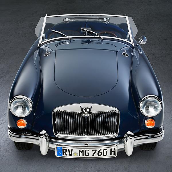 MGA 1600 Roadster Front Ein blauer Oldtimer, ein MG, steht aus der Vogelperspektive auf einem glatten Untergrund. Die Motorhaube ist glänzend, und die Scheinwerfer leuchten. Die Windschutzscheibe ist klar, und das Fahrzeug hat keine Dachabdeckung, was einen offenen Eindruck vermittelt.
