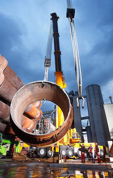 Reparaturarbeiten Ein Kran hebt ein großes Rohr an einer Baustelle in einer Industrieumgebung. Im Hintergrund sind weitere Baugeräte und ein bewölkter Himmel sichtbar. Arbeiter stehen in der Nähe und beobachten den Vorgang.