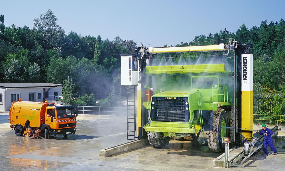 Waschanlage fuer Schwerlastkraftwagen Ein grüner Großtraktor wird von einer automatischen Waschanlage gereinigt. Neben der Waschanlage steht ein orangefarbener Reinigungswagen. Wasser spritzt überall, während ein Arbeiter in blauer Arbeitskleidung zuschaut. Im Hintergrund sind Bäume und eine Gebäudeansicht sichtbar.