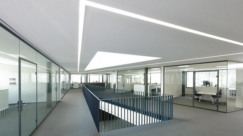 Schmieder Gebaeude Moderne Bürolandschaft mit Glaswänden und einem offenen Bereich. Helle, minimalistische Gestaltung mit natürlichem Licht, das durch ein großes Dachfenster strömt. Ein erhöhter Gang führt zwischen den Büroräumen. Der Boden ist mit grauem Teppich ausgelegt.