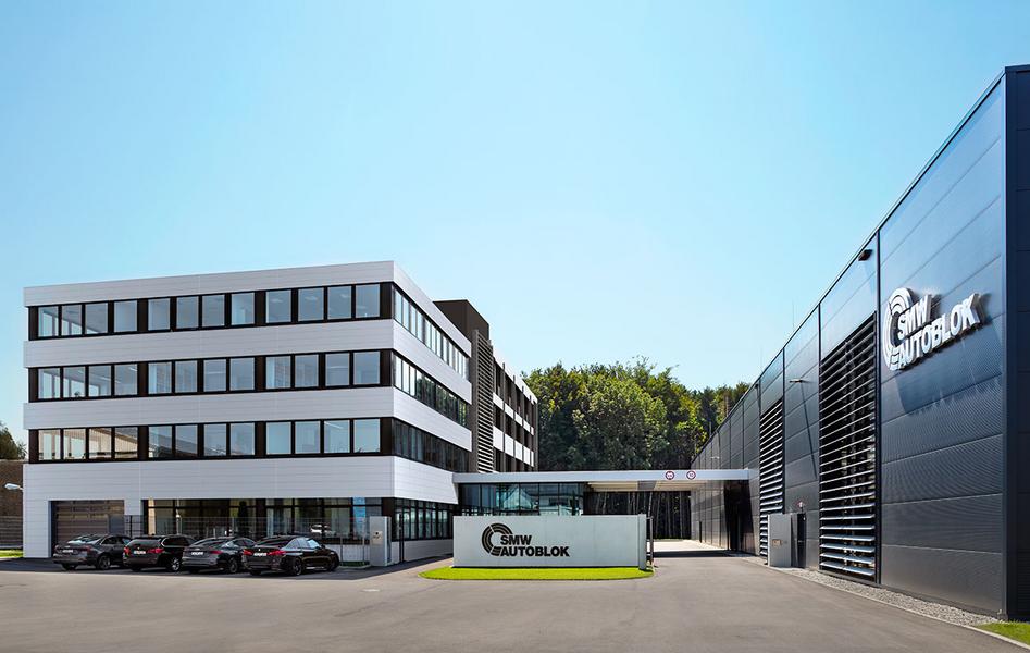 Verwaltungs und Produktionsgebaeude Moderne Unternehmensgebäude mit zwei Abschnitten, einem mehrstöckigen weißen Gebäude und einem schwarzen, modernen Anbau. Im Vordergrund steht ein Schild mit dem Firmenlogo. Vor dem Gebäude parken mehrere Autos. Der Himmel ist klar und blau.