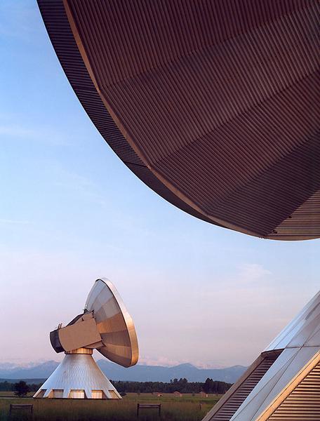 Erdfunkstation Zwei große Parabolantennen stehen auf einem offenen Feld. Die vordere Antenne ist leicht geneigt, während die hintere Antenne schräg im Hintergrund sichtbar ist. Der Himmel ist klar und zeigt sanfte Farben des Sonnenuntergangs.