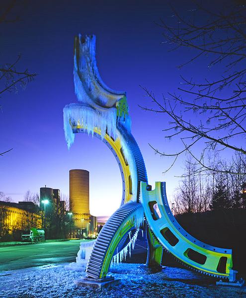Skulptur vor einem Zementwerk Eine große, bunte Skulptur in Form eines Zahnrads, das mit Eiszapfen bedeckt ist. Im Hintergrund sind Industriegebäude und ein Kühlturm sichtbar. Der Himmel ist in tiefen Blau- und Lilatönen erleuchtet, was eine kalte, aber beeindruckende Abendstimmung vermittelt.