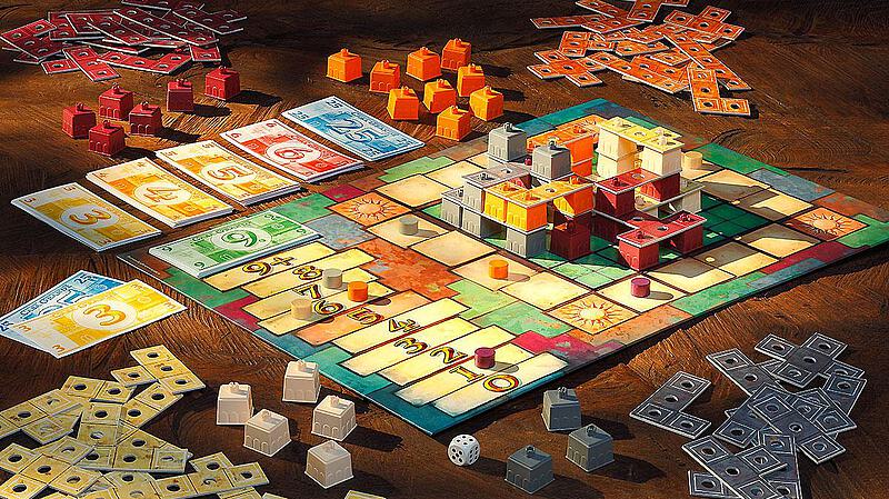 Ein Tisch mit einem aufgeräumten Brettspiel. In der Mitte liegt das Spielbrett mit bunten Spielfiguren und Karten. Rund um das Brett sind Stapel aus farbigen Spielsteinen und Geldscheinen verteilt. Ein Würfel liegt ebenfalls auf dem Tisch.