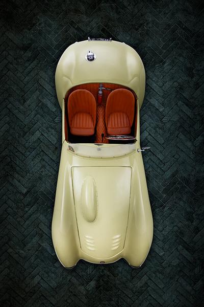 FIAT 500 Graziani MM Draufsicht Ein cremefarbener Oldtimer mit roten Ledersitzen, aus der Vogelperspektive betrachtet. Der Wagen hat eine geschwungene Form und edle Details. Der Hintergrund ist aus dunklen, geflammten Platten gefertigt.