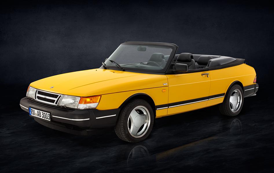 Saab 900 i Cabriolet Front Ein gelber Sportwagen-Cabrio, Modell Saab, steht seitlich auf einer dunklen, einfarbigen Oberfläche. Das Auto hat schwarze Details und ein offenes Verdeck, sichtbar sind die beiden vorderen Sitze im Innenraum.