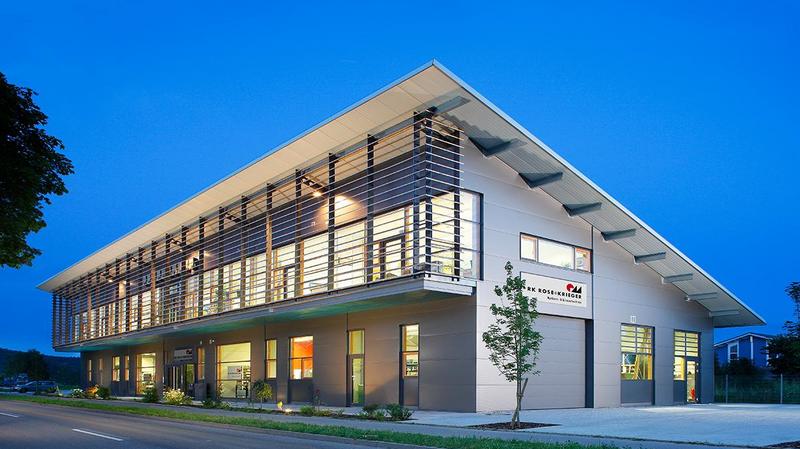 Produktionsgebaeude Moderner, zweigeschossiger Gebäude in der Dämmerung. Große Fenster und eine klare, rechteckige Struktur. Vor dem Gebäude ist ein kleiner Baum sowie eine asphaltierte Straße zu sehen. Die Fassade hat vertikale Holzleisten und eine klare Beschriftung am Eingang.