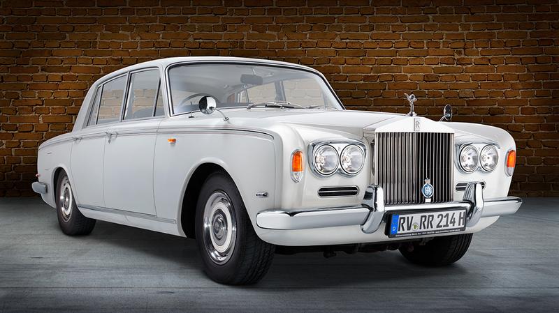 Rolls Royce Silver Shadow Front Ein klassisches weißes Auto, ein Rolls-Royce, steht vor einer Backsteinmauer. Die Front weist markante Scheinwerfer und das bekannte Kühleremblem auf. Das Fahrzeug hat eine elegante und luxuriöse Erscheinung.