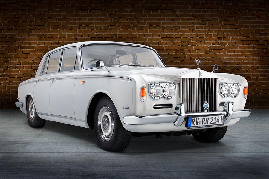Rolls Royce Silver Shadow Front Ein klassisches weißes Auto, ein Rolls-Royce, steht vor einer Backsteinmauer. Die Front weist markante Scheinwerfer und das bekannte Kühleremblem auf. Das Fahrzeug hat eine elegante und luxuriöse Erscheinung.