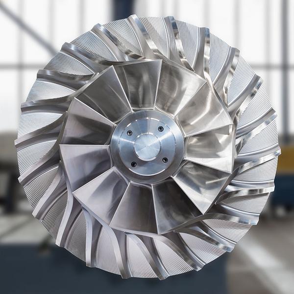 Turbinenlaufrad Detailansicht eines Turbinenrotors aus Metall. Die Rotorblätter sind symmetrisch angeordnet und haben eine geschwungene Form. Im Hintergrund sind unscharfe Werkstattelemente zu erkennen.
