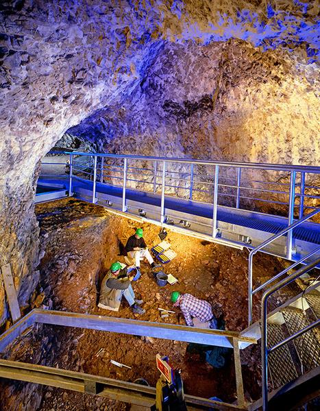 Hohle Fels Schelklingen Eine Gruppe von Arbeitern mit grünen Helmen gräbt in einem Erdloch in einer Höhle. Über ihnen verläuft eine Metallbrücke, und die Wände der Höhle sind aus Stein. Die Beleuchtung erzeugt ein blaues Licht, das den Raum akzentuiert.