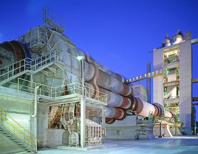 Drehofen zur Zementproduktion Industrieanlage bei Nacht, mit einem großen rotierenden Kamin, der in einer modernen Betonanlage eingebaut ist. Säulen und Treppenverläufe sind sichtbar, sowie verschiedene Maschinen und Förderanlagen. Die Umgebung ist in kühlem Blau beleuchtet.