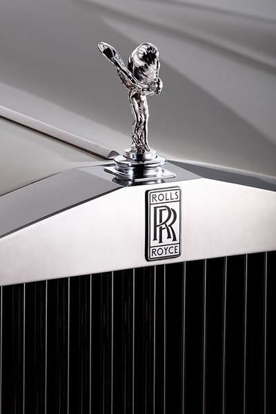 Rolls Royce Silver Shadow Emily Eine Nahaufnahme des Kühlergrills eines Rolls-Royce mit dem emblematischen "Spirit of Ecstasy" (eine stilisierte Figur) und dem Rolls-Royce-Logo. Der Grill ist glänzend und hat vertikale Streifen.