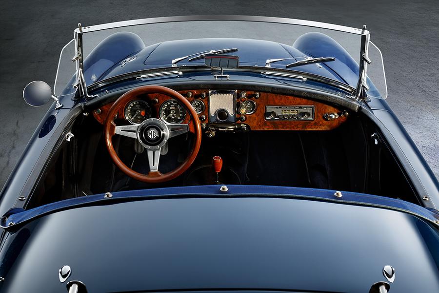 MGA 1600 Roadster Cockpit Eine Innenansicht eines klassischen Autos, das von oben fotografiert wurde. Der Blick zeigt das Lenkrad aus Holz, das Armaturenbrett mit verschiedenen Instrumenten und einem Radio. Die Sitze sind schwarz und der Fahrzeugboden ist gut sichtbar.