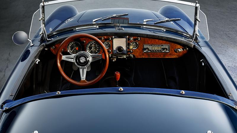 MGA 1600 Roadster Cockpit Eine Innenansicht eines klassischen Autos, das von oben fotografiert wurde. Der Blick zeigt das Lenkrad aus Holz, das Armaturenbrett mit verschiedenen Instrumenten und einem Radio. Die Sitze sind schwarz und der Fahrzeugboden ist gut sichtbar.