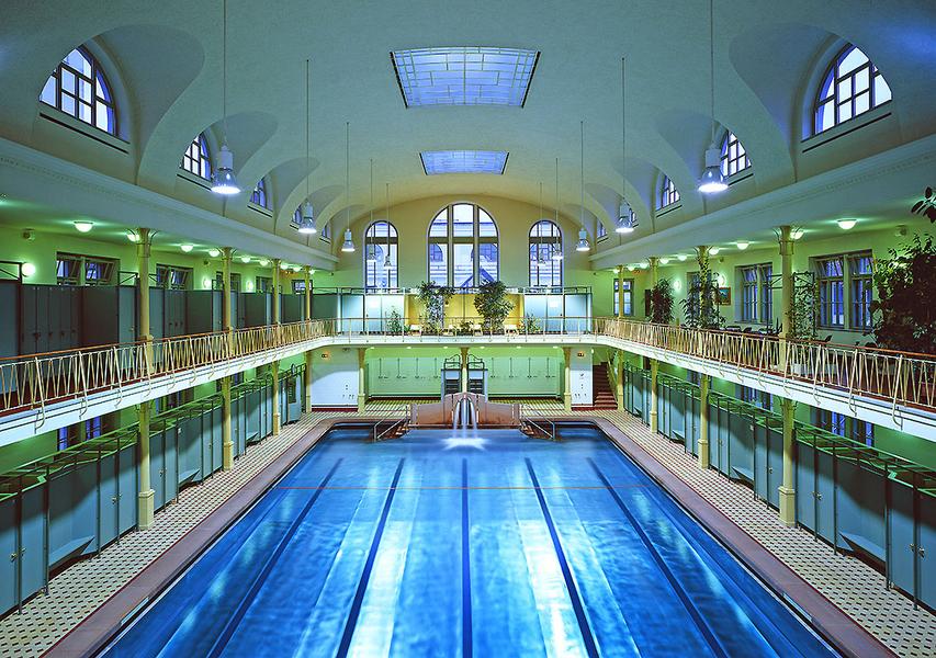 Historische Badeanstalt Ein großer, eleganter Innenraum eines Schwimmbads mit einem blauen Schwimmbecken in der Mitte. Die Wände sind mit Fenstern und Pflanzen geschmückt, und große, gewölbte Decken sorgen für eine helle, offene Atmosphäre.