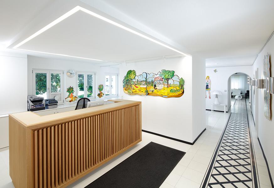 Empfang Eine modern gestaltete Empfangshalle mit einem hölzernen Empfangstresen. An der Wand hängt ein farbenfrohes Wandbild einer Landschaft. Der Boden ist hell gefliest und im Hintergrund sind weitere Räume sichtbar.