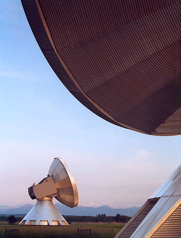 Erdfunkstation Zwei große Parabolantennen stehen auf einem offenen Feld. Die vordere Antenne ist leicht geneigt, während die hintere Antenne schräg im Hintergrund sichtbar ist. Der Himmel ist klar und zeigt sanfte Farben des Sonnenuntergangs.