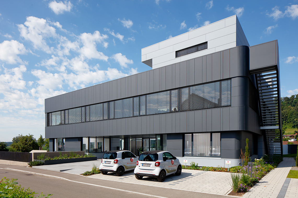 Schmieder Gebaeude Modernes, mehrstöckiges Gebäude mit großen Fenstern und grauer Fassadenverkleidung. Vor dem Gebäude stehen zwei kleine Autos auf einem gepflasterten Parkplatz. Im Hintergrund sind Bäume und ein blauer Himmel mit einigen Wolken sichtbar.