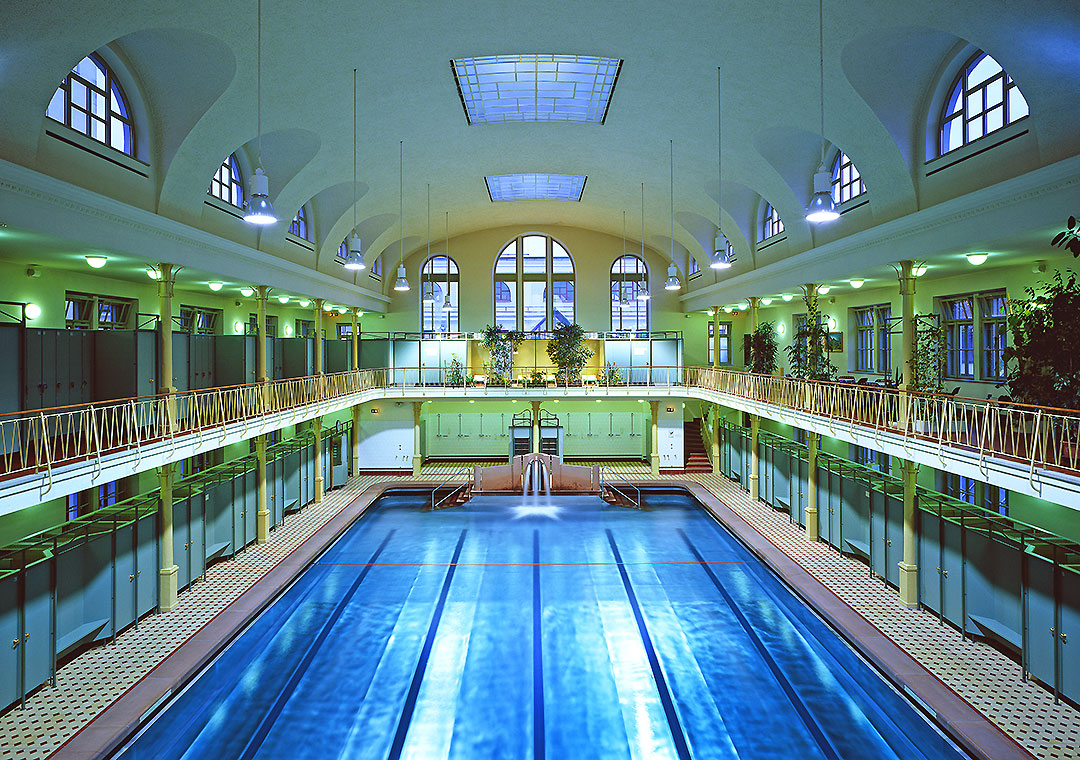 Historische Badeanstalt Ein großer, eleganter Innenraum eines Schwimmbads mit einem blauen Schwimmbecken in der Mitte. Die Wände sind mit Fenstern und Pflanzen geschmückt, und große, gewölbte Decken sorgen für eine helle, offene Atmosphäre.