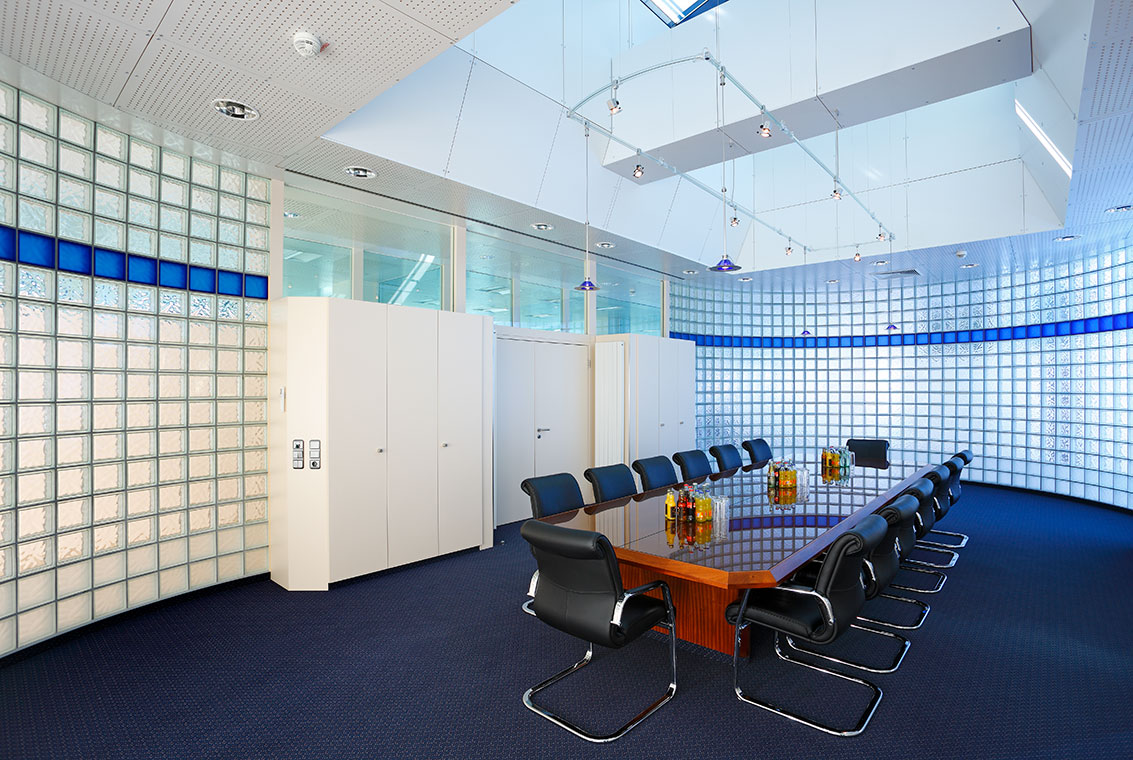 Konferenzraum Ein moderner Besprechungsraum mit einem großen, rechteckigen Tisch in der Mitte, umgeben von schwarzen Stühlen. Die Wände sind mit transparenten Glasbausteinen dekoriert, und an der Decke befinden sich Fenster, die Tageslicht hereinlassen.