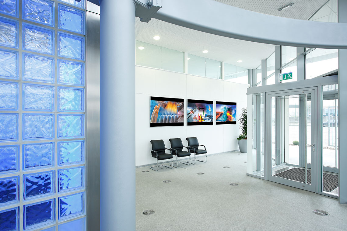 Empfang Ruhiger Empfangsbereich mit Glaswand und mehreren Sitzplätzen. An der Wand hängen drei Bilder mit lebhaften Farben. Die Eingangstür ist aus Glas, und in der Ecke steht eine Pflanze.