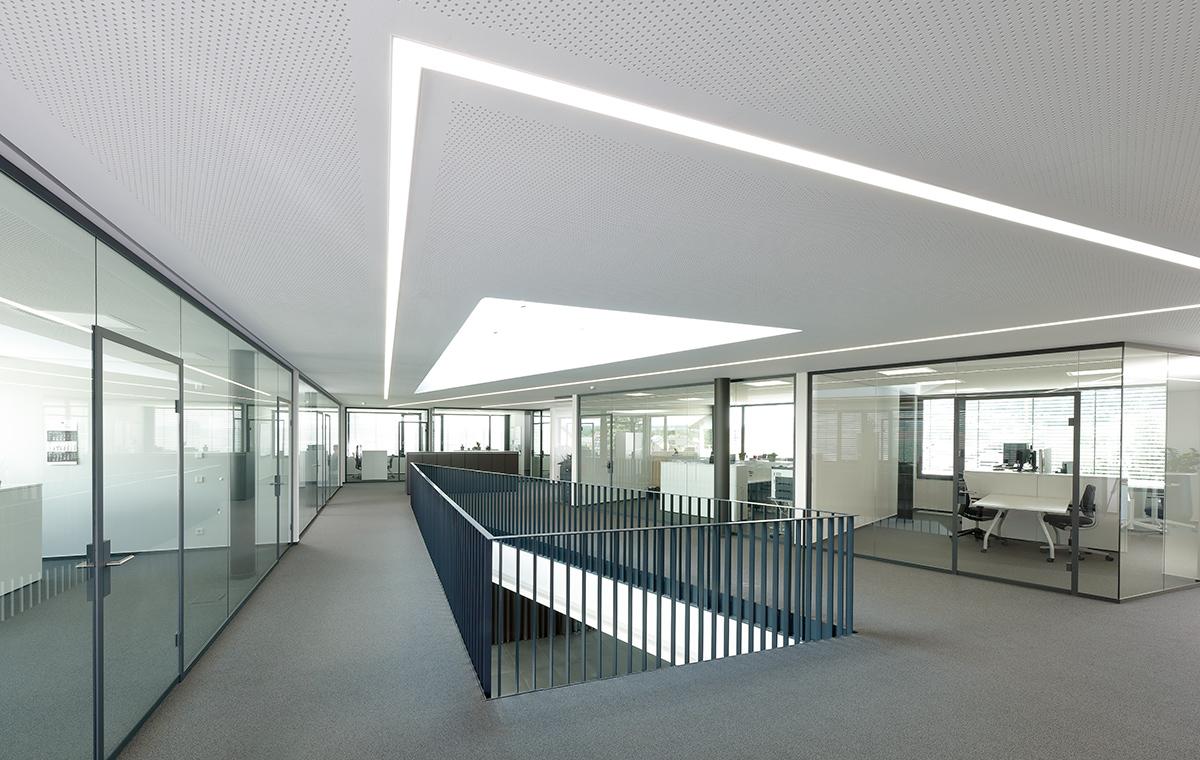 Schmieder Gebaeude Moderne Bürolandschaft mit Glaswänden und einem offenen Bereich. Helle, minimalistische Gestaltung mit natürlichem Licht, das durch ein großes Dachfenster strömt. Ein erhöhter Gang führt zwischen den Büroräumen. Der Boden ist mit grauem Teppich ausgelegt.