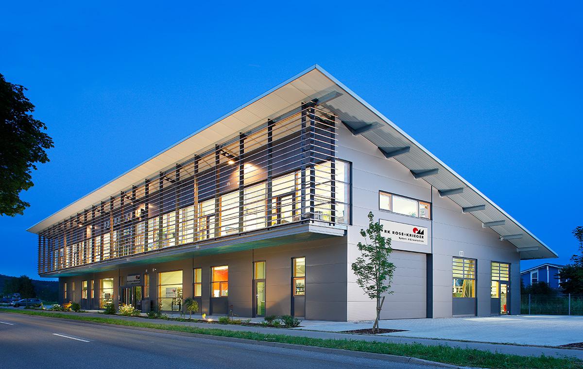 Produktionsgebaeude Moderner, zweigeschossiger Gebäude in der Dämmerung. Große Fenster und eine klare, rechteckige Struktur. Vor dem Gebäude ist ein kleiner Baum sowie eine asphaltierte Straße zu sehen. Die Fassade hat vertikale Holzleisten und eine klare Beschriftung am Eingang.