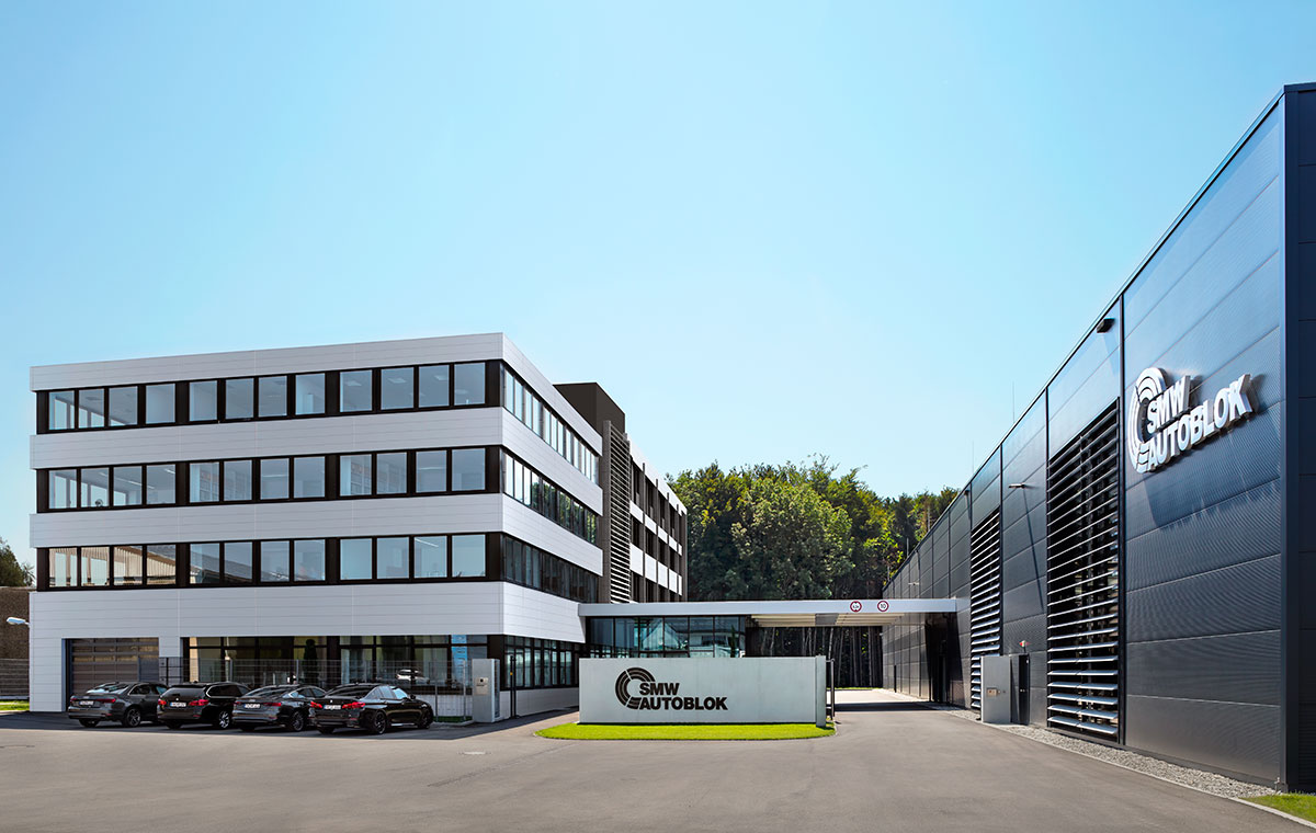 Verwaltungs und Produktionsgebaeude Moderne Unternehmensgebäude mit zwei Abschnitten, einem mehrstöckigen weißen Gebäude und einem schwarzen, modernen Anbau. Im Vordergrund steht ein Schild mit dem Firmenlogo. Vor dem Gebäude parken mehrere Autos. Der Himmel ist klar und blau.