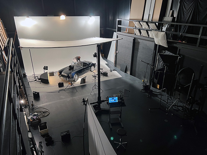 Studio Ein Blick auf ein Fotostudio mit einem schwarzen Auto auf einem weißen Hintergrund. Ein Fotograf arbeitet am Auto, während mehrere Scheinwerfer und Kameras aufgestellt sind. Der Raum ist dunkel, und es sind verschiedene Studioeinrichtungen sichtbar.