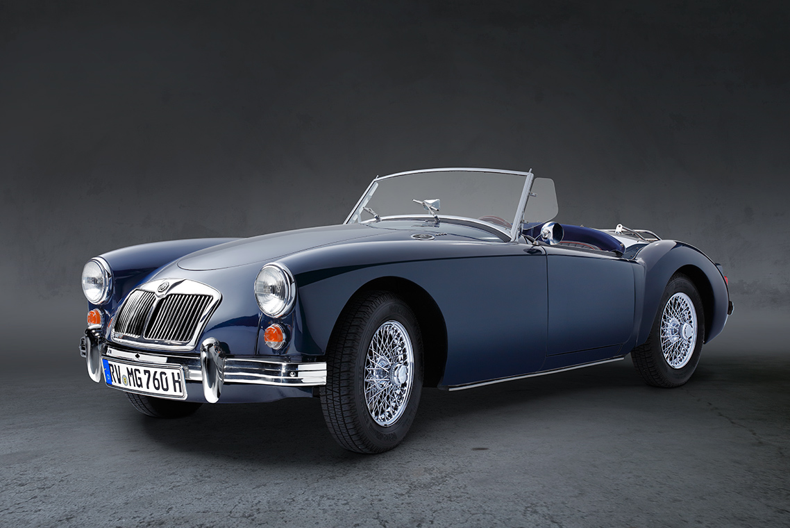 MGA 1600 Roadster Front Ein blauer Oldtimer-Cabriolet mit offenem Verdeck steht auf einer neutralen, grauen Fläche. Die glänzenden Felgen und der Charme des klassischen Designs heben sich von der dunklen Hintergrundfarbe ab.