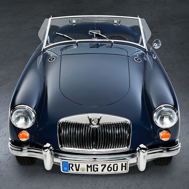 Ein blauer Oldtimer, ein MG, steht aus der Vogelperspektive auf einem glatten Untergrund. Die Motorhaube ist glänzend, und die Scheinwerfer leuchten. Die Windschutzscheibe ist klar, und das Fahrzeug hat keine Dachabdeckung, was einen offenen Eindruck vermittelt.