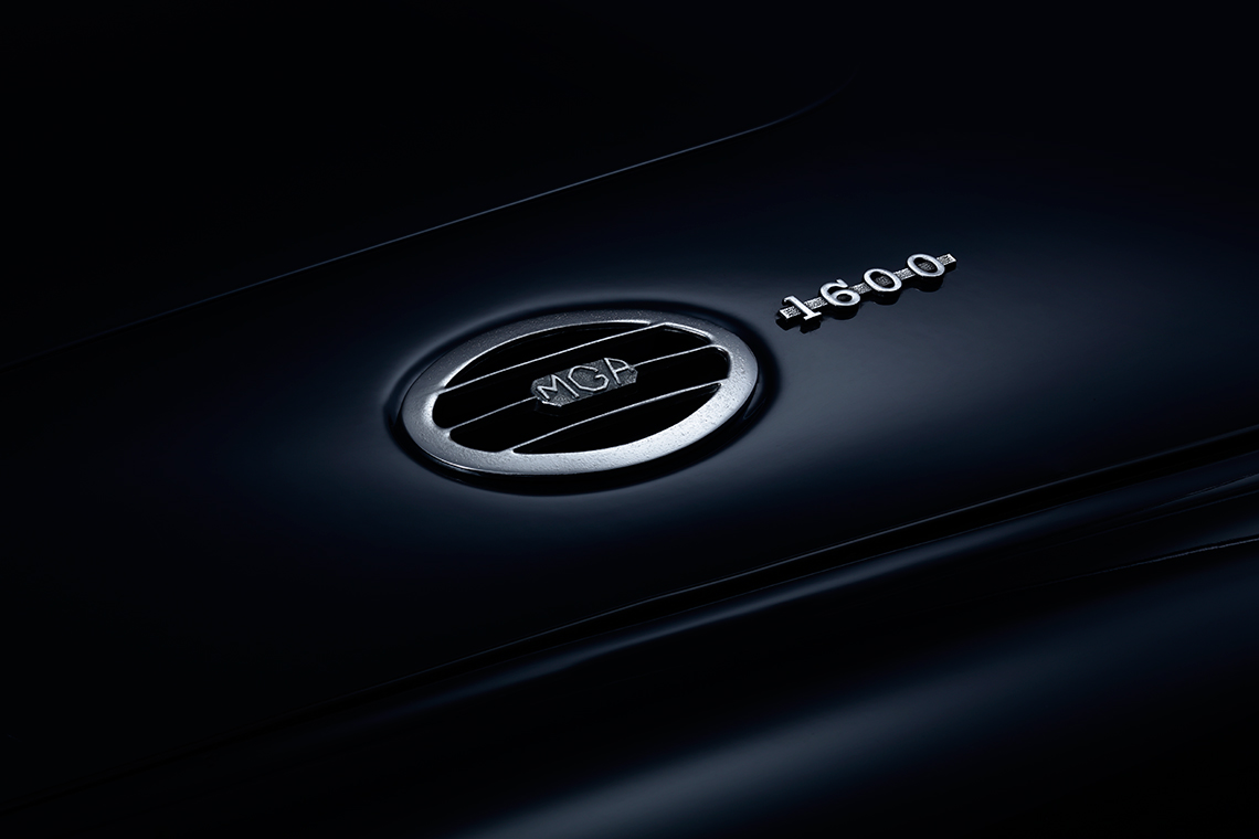 MGA 1600 Roadster Hutze Ein schimmerndes Logo und die Modellbezeichnung "MGA 1600" auf einer dunklen Fahrzeugoberfläche. Das Logo hat eine runde Form mit horizontalen Streifen und ist in silberner Farbe gehalten.
