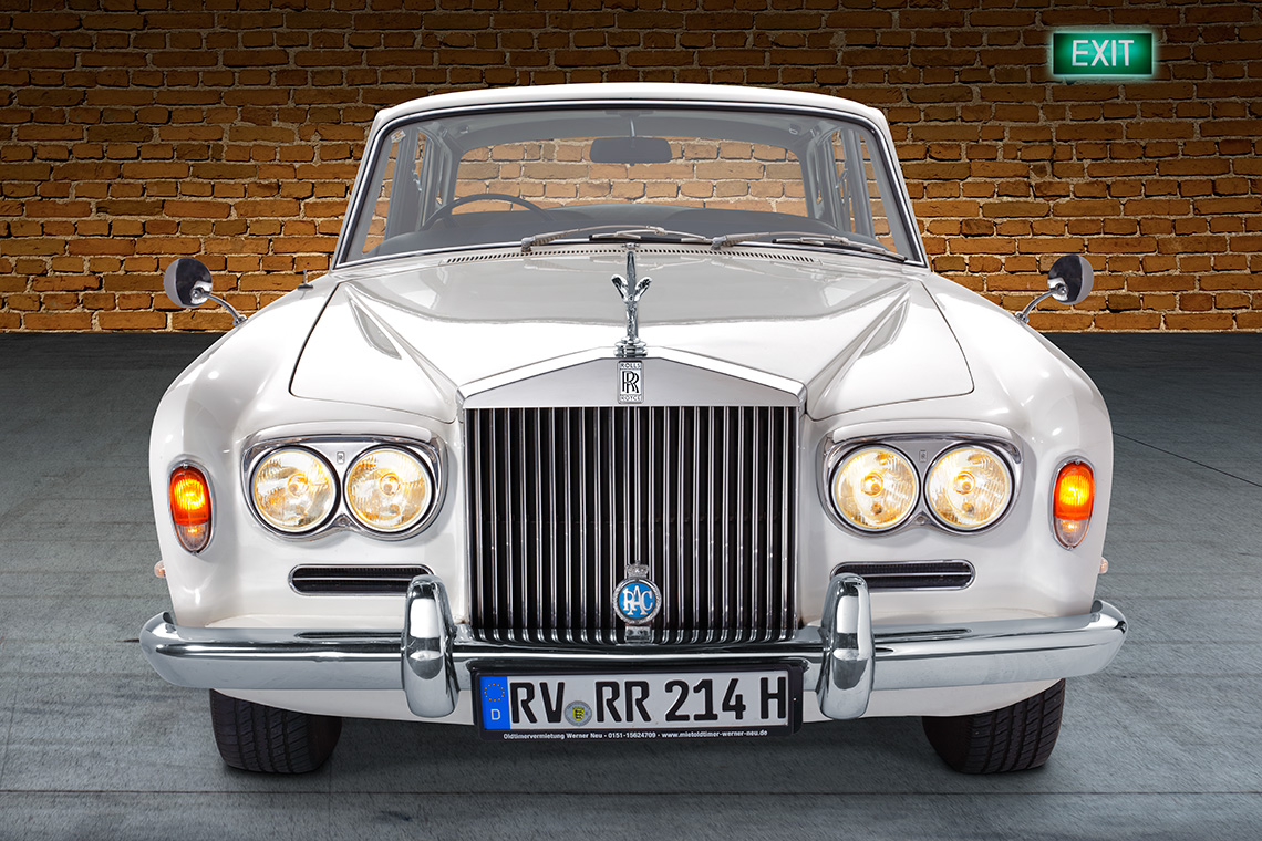 Rolls Royce Silver Shadow Front Ein klassischer, weißer Rolls-Royce mit markanter Kühlergrill und runden Scheinwerfern. Das Fahrzeug steht vor einer Backsteinwand. Ein grünes Ausstiegsschild ist im Hintergrund sichtbar.